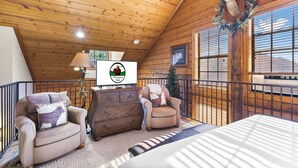 Interior acogedor de una cabaña con paredes de madera, techo con vigas a la vista, chimenea, cabeza de ciervo montada y un televisor que muestra un logotipo.