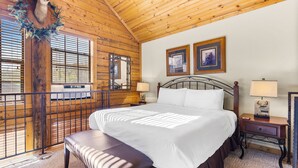 Quarto com cama grande, teto de madeira, duas imagens emolduradas, decoração de cabeça de veado e uma janela com persianas.