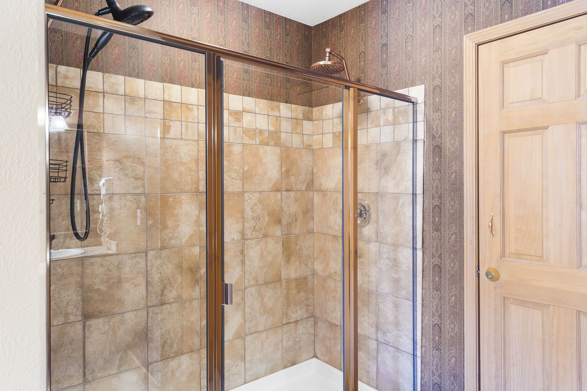 A glass-enclosed shower with beige tiles, a wooden door, and a patterned wall.