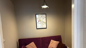 Un salon avec un canapé violet, deux coussins, une œuvre d’art abstraite encadrée accrochée au mur et un luminaire au plafond.