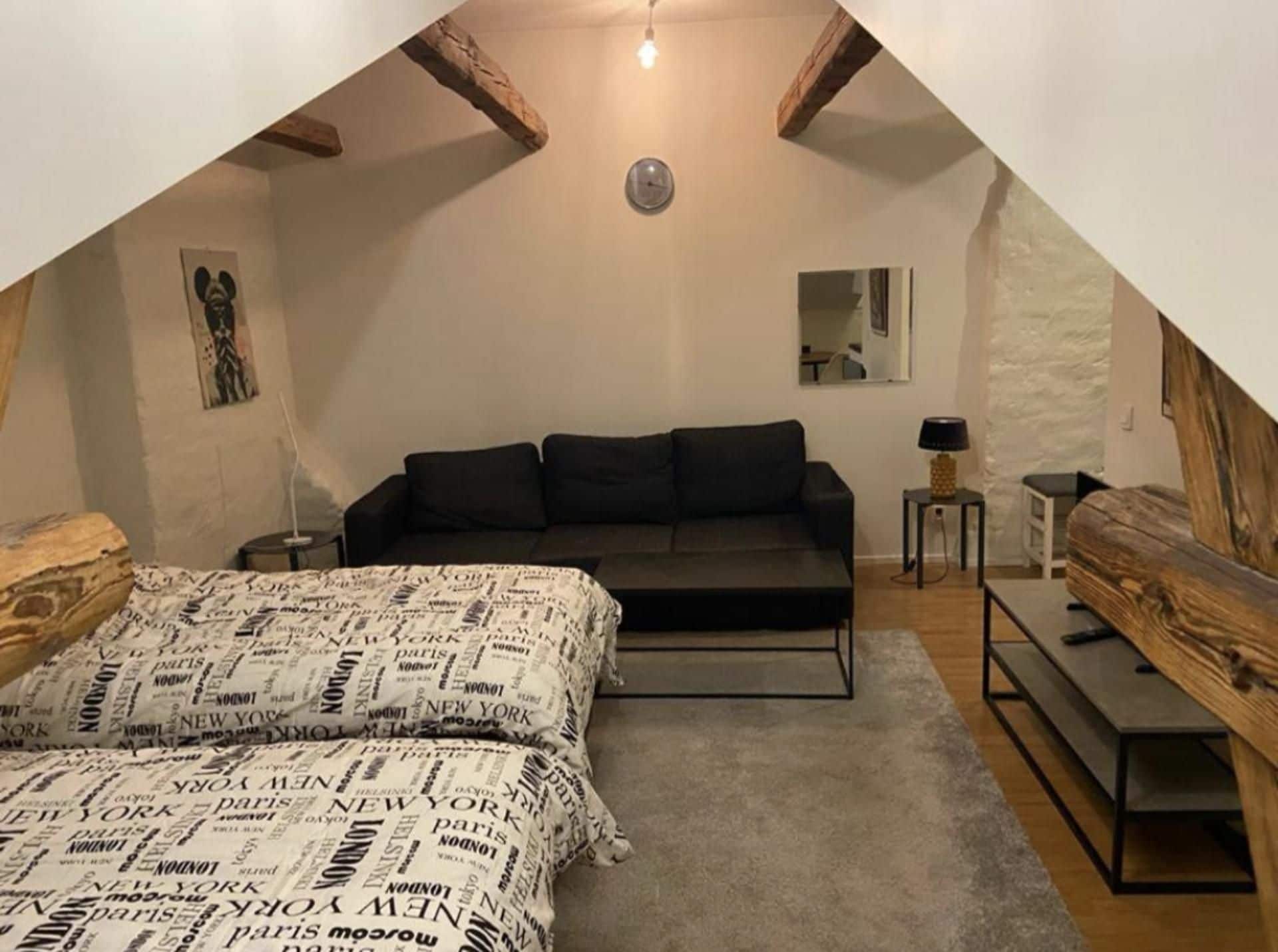 Un dormitorio con techo inclinado, una cama con colcha estampada, un sofá oscuro, una mesita de noche con una lámpara y un espejo en la pared.