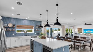 Una cucina moderna con isola centrale, pensili bianchi e zoccolo blu. C'è un'area pranzo con tavolo e sedie, e un'ampia finestra che offre vista sull'esterno.