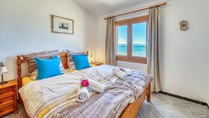 A neatly made bed with a view of the sea through a window, a wooden headboard, and a small bedside table with a lamp. - Villa in front of the sea Casa Sa Nau in Mallorca (Arta)