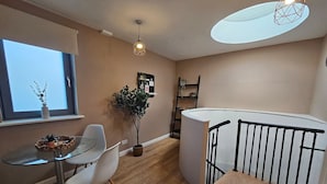 A modern living room with a glass dining table, white chairs, a potted plant, a black metal railing, and a skylight.