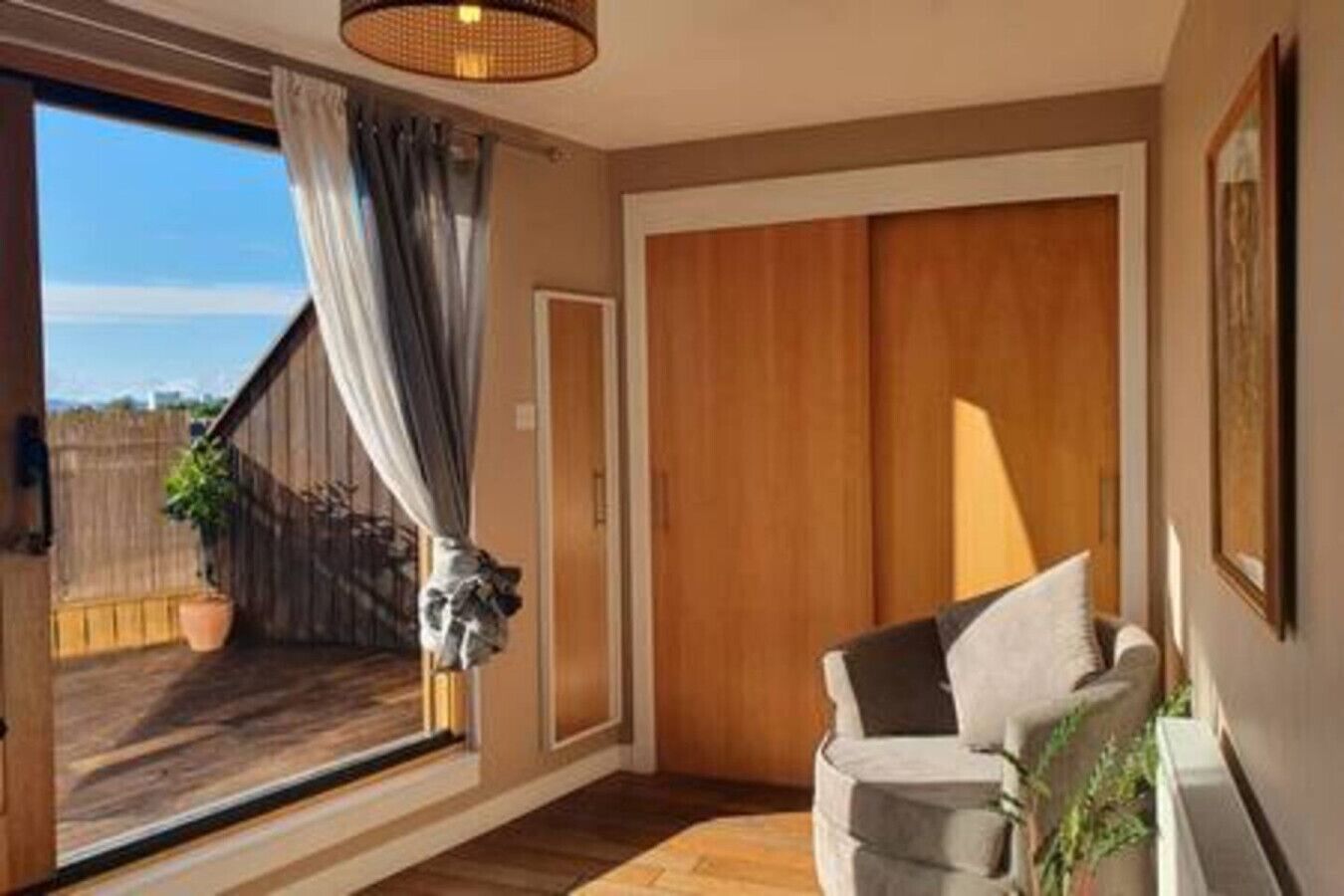 A room with a sliding glass door leading to a balcony, a wooden floor, a sofa with cushions, and a hanging light fixture.