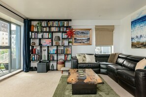 A modern living room with a leather sofa, a bookshelf, a coffee table, and a large window.