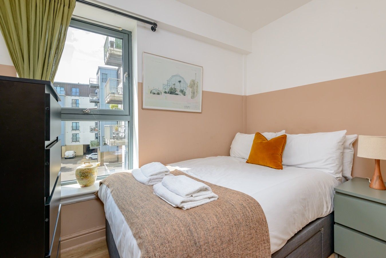 A hotel room with a bed, a window with a view of buildings, a framed picture on the wall, and a lamp on the nightstand.