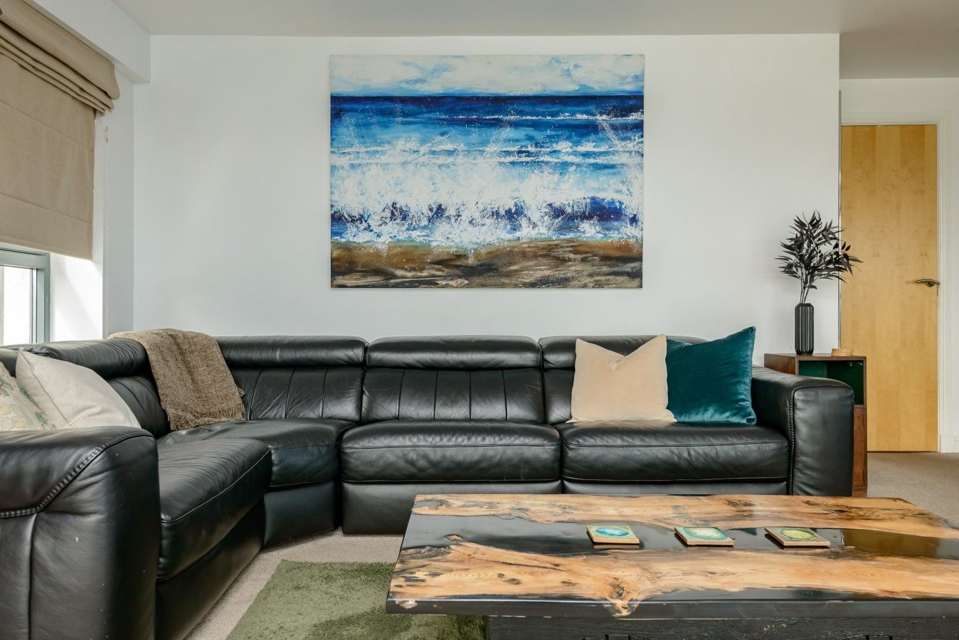 A living room with a black leather sofa, a coffee table with a wooden top, and a large painting of a seascape on the wall.