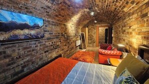 A room with exposed brick walls, a bed with orange bedding, a sofa with red cushions, and a painting on the wall. - The Armoury: Stylish WW1 Bunker (North Queensferry)