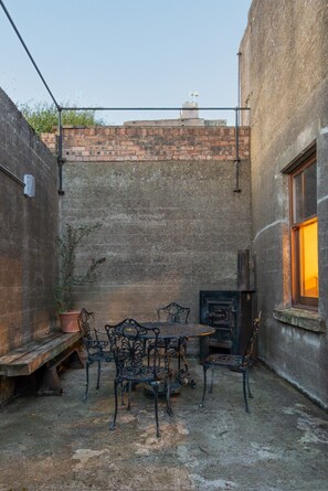 A courtyard with a brick wall, a metal table with chairs, a potted plant, and a window with warm light.