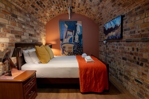 A hotel room with a brick wall, a bed with pillows, a bedside table, and a painting on the wall.