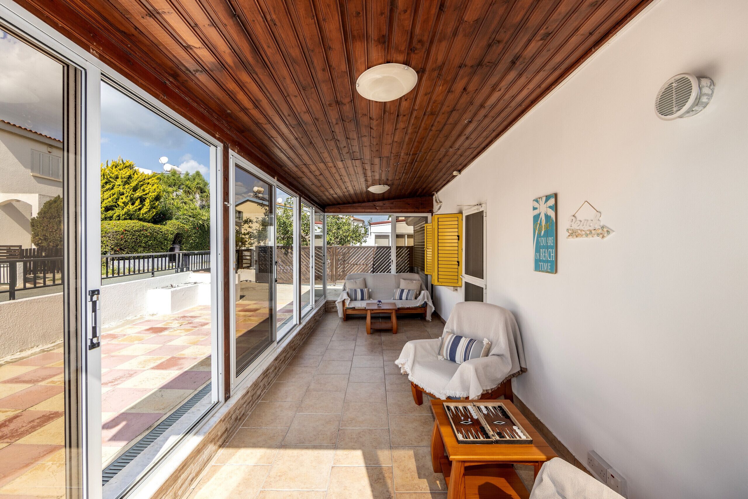 A covered patio with a wooden ceiling, sliding glass doors, a sofa, a table, and a chair.