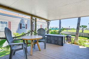 Eine überdachte Terrasse im Freien mit einem runden Tisch und zwei Stühlen, einem Grill und Blick auf eine Wohngegend mit Häusern und Palmen.