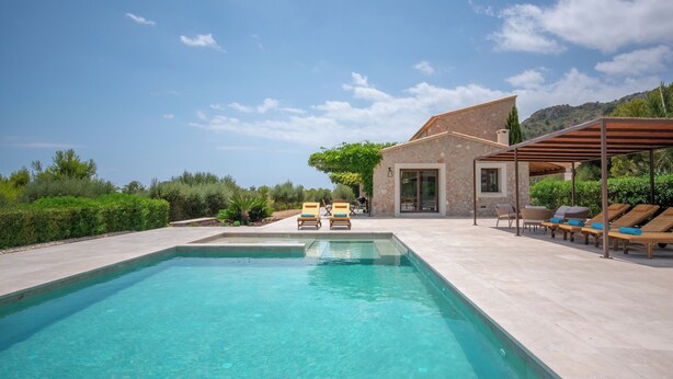 Una piscina rectangular con agua azul cristalina, rodeada por una terraza de baldosas de color claro. Hay dos sillones de jardín con cojines y una estructura de pérgola de madera.