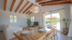 Un salón moderno con una mesa de comedor de vidrio, vigas de madera, un sofá, un televisor y vistas al exterior.