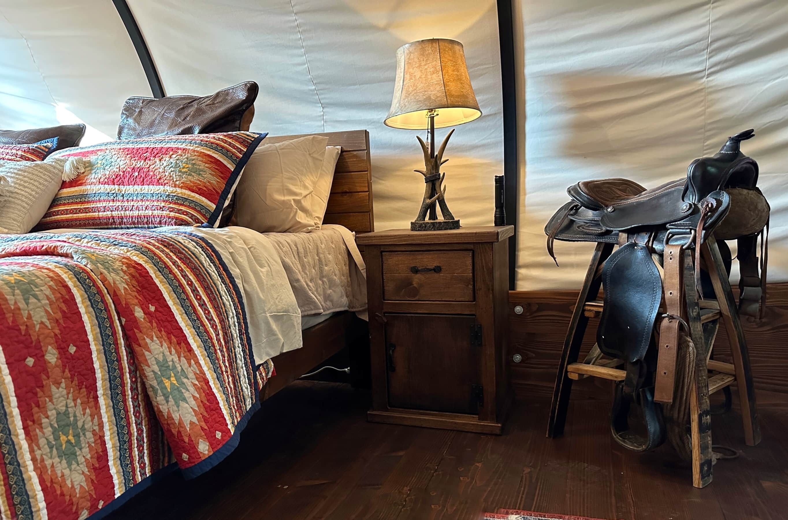 A cozy tented room with a bed, a wooden nightstand with a lamp, and a saddle chair.
