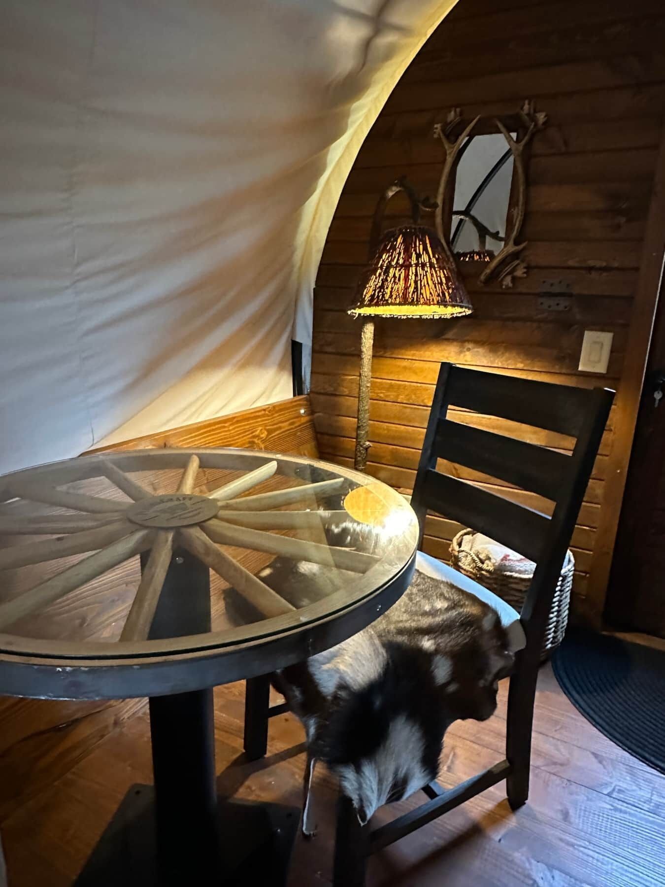 A round glass table with a wooden base, a woven lamp, a wooden chair, and a rustic wall with a mirror.