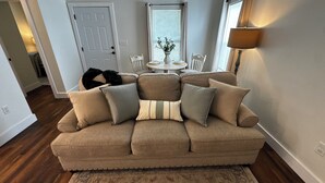 Ein Wohnzimmer mit einem beigen Sofa, einem gemusterten Zierkissen, einem kleinen runden Tisch mit einer Vase und einer Lampe.