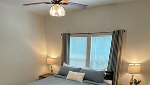 Ein Schlafzimmer mit einem Bett, Deckenventilator, zwei Nachttischlampen, einem Nachttisch, einem Fenster mit Vorhängen und einem Teppich.