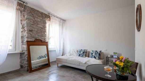 A room with a stone wall, a bed with patterned pillows, a small table with a vase of flowers, and a mirror on the wall.