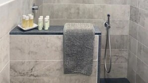 A small, tiled shower room with a handheld showerhead, a towel rack, and a shelf with toiletries.