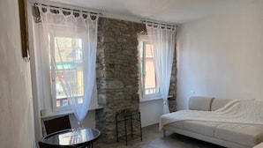 A small, cozy room with a stone wall, a white sofa, a round table with chairs, and two windows with sheer curtains.