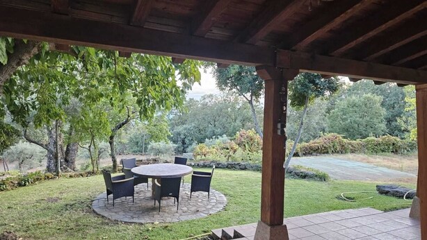 A covered patio with a round table and chairs, surrounded by a garden with trees and a lawn. - Casa Rural (Candeleda)