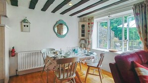 A dining area with a table set for four, wooden chairs, a red sofa, a window with a view of greenery, a white radiator, and a small birdhouse on the wall. - Discover a cosy two-bedroom cottage on the Colmer Estate in Ivybridge. (Ivybridge)