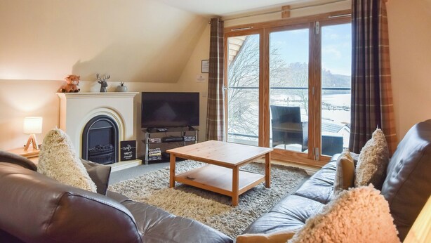 A living room with a fireplace, a television, a sofa, and a view of a mountainous landscape through large windows. - 4 bedroom accommodation in Balmaha (Balmaha)