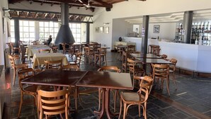 A spacious restaurant with high ceilings, wooden beams, and a central black column. Tables and chairs are arranged for dining.