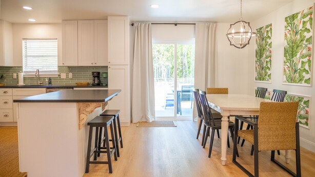 Une cuisine moderne dotée d’un îlot central, de meubles blancs et d’un coin repas avec des chaises en osier et une table blanche.
