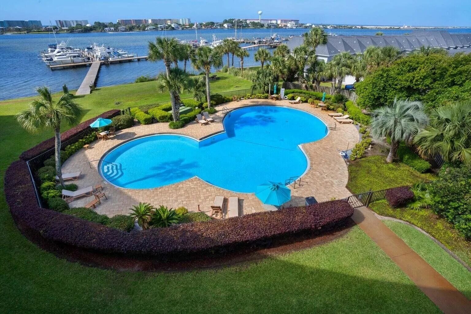 A large, curved swimming pool with multiple lounge areas, surrounded by palm trees and a view of the marina.
