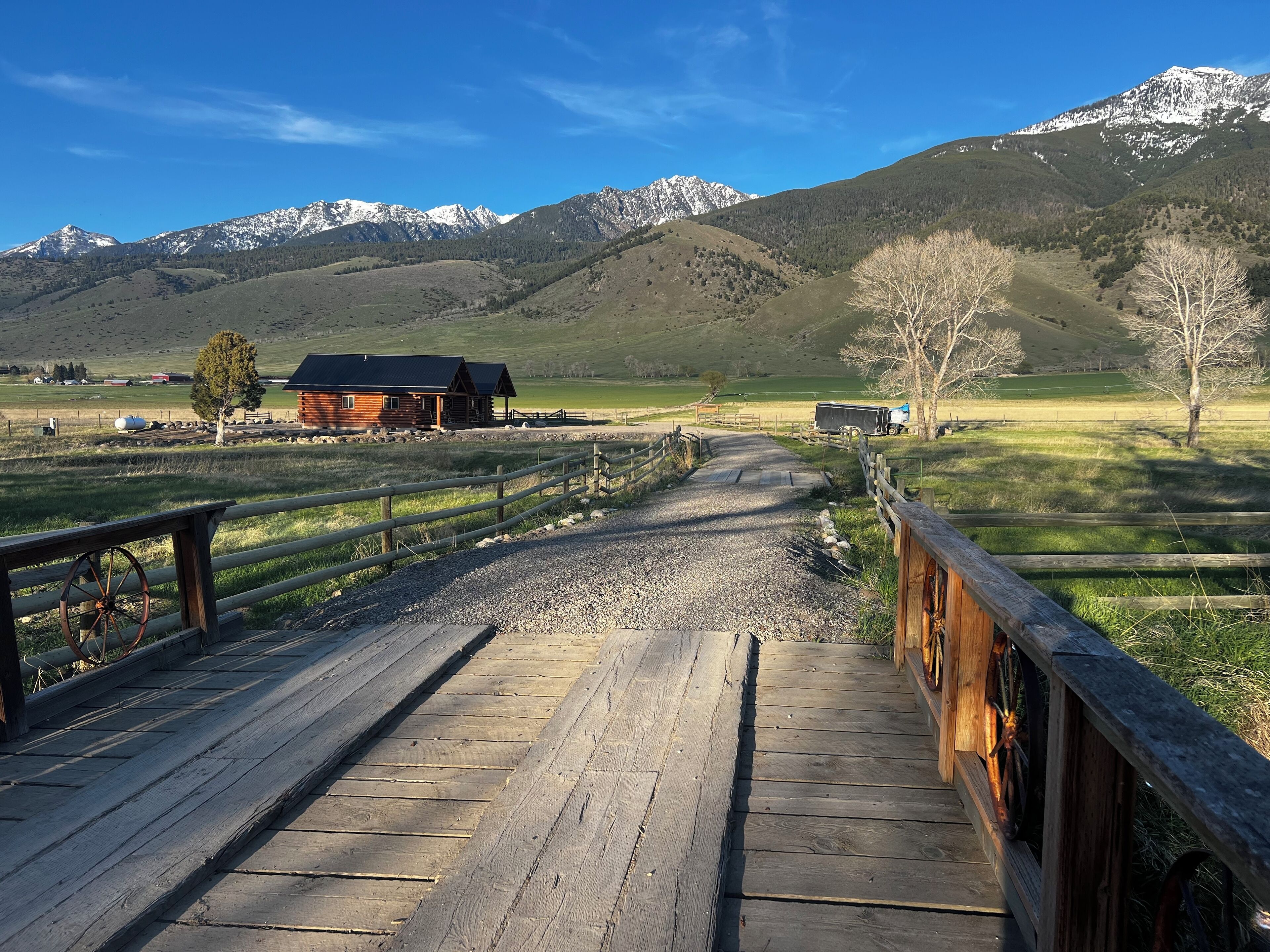 A wooden bridge leading to a cabin in a mountainous area with snow-capped peaks in the background.