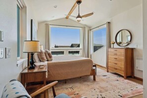 Un dormitorio con cama, cómoda, escritorio con lámpara, ventilador de techo y ventanas amplias que ofrecen vistas.