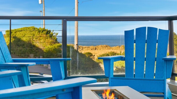 A view from a room with large windows overlooking a beach, featuring blue Adirondack chairs and a small fire table. - Light House by Manzanita Beach Getaway: High-End w/ Ocean & Neahkahnie Views (Manzanita)