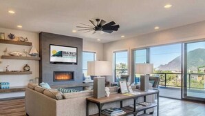 A modern living room with a fireplace, a large window with a mountain view, and a ceiling fan. - Light House by Manzanita Beach Getaway: High-End w/ Ocean & Neahkahnie Views (Manzanita)