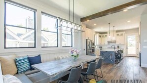 A modern kitchen with a central island, large windows, and a dining area with a long table and chairs. - Light House by Manzanita Beach Getaway: High-End w/ Ocean & Neahkahnie Views (Manzanita)