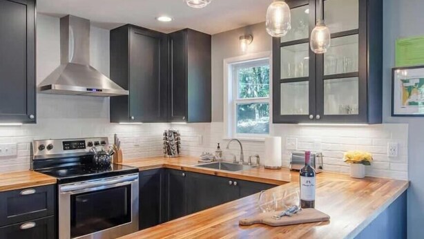 Une cuisine moderne avec des meubles de couleur sombre, une hotte en acier inoxydable, un plan de travail en bois et des luminaires suspendus.