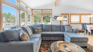 A modern living room with a large sectional sofa, a glass coffee table, and a dining area with a table and chairs. - Coastal View by Manzanita Beach Getaway: Ocean Views, Dog-Friendly & Great Deck (Neahkahnie)