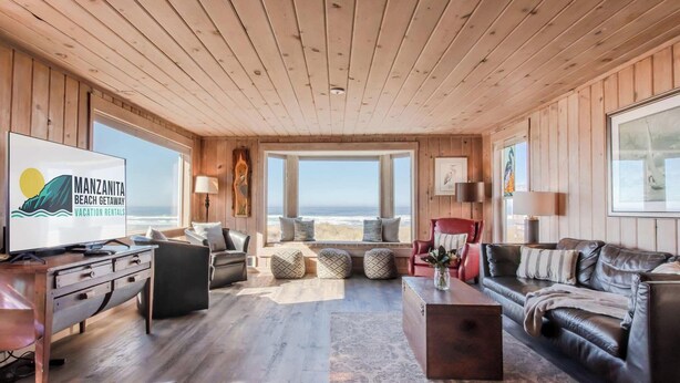 Een ruime woonkamer met houten wanden en plafond, een groot raam met uitzicht op het strand, een flatscreen-tv en een gezellige zithoek met een salontafel.