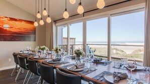 Een eetruimte met een lange tafel gedekt voor zes personen, grote ramen met uitzicht op het strand, en hangende lampen.