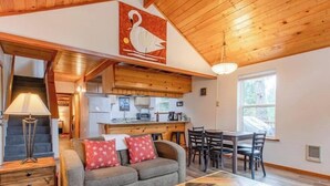 L’intérieur chaleureux d’un chalet avec un plafond en bois, un coin repas, une cuisine et un salon doté d’un canapé et de coussins.