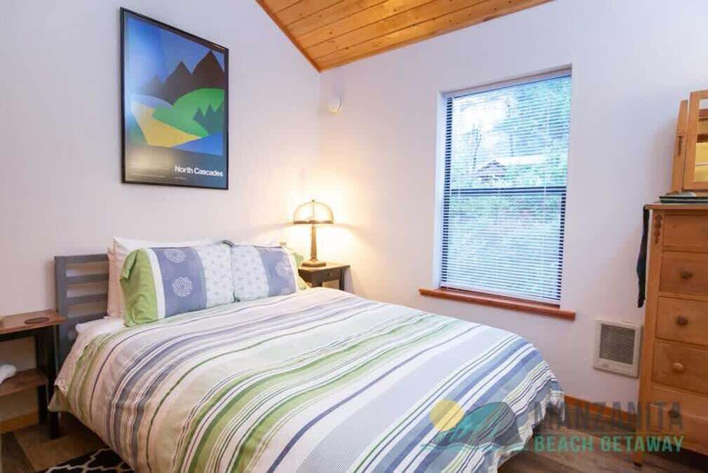A bedroom with a bed, a nightstand, a lamp, a window with blinds, a dresser, and a framed picture on the wall.
