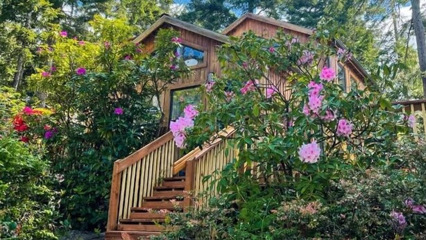Un escalier en bois mène à une maison entourée d’une végétation luxuriante et de fleurs roses.