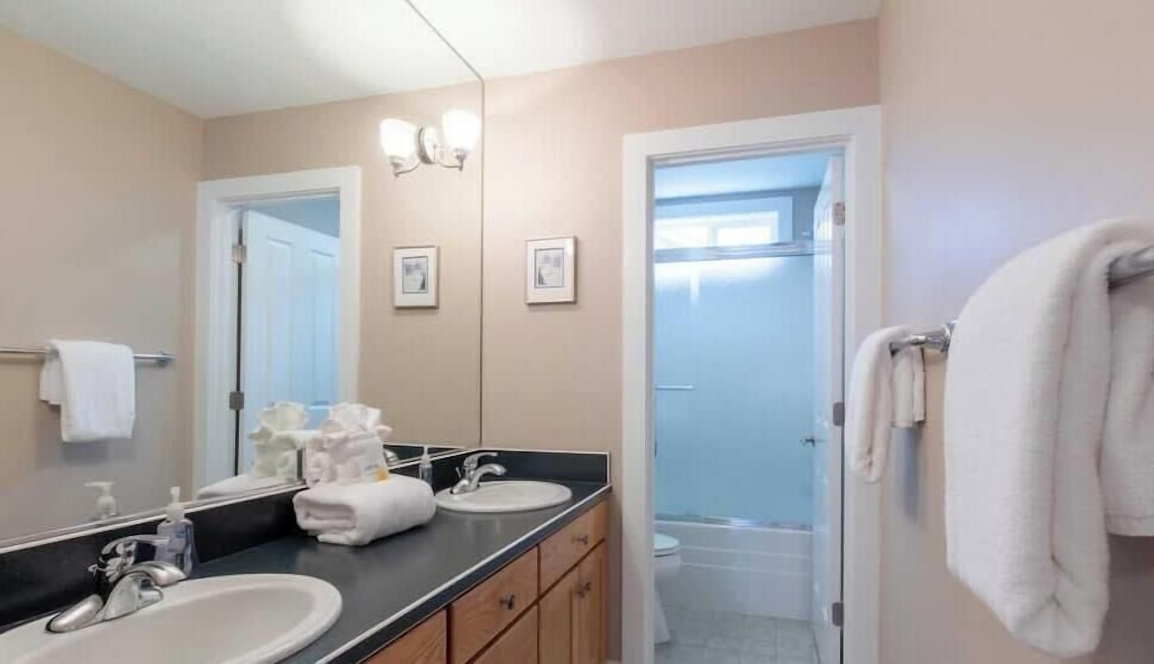 Een badkamer met twee wastafels, een spiegel en een toilet. Handdoeken hangen aan de muur en er is een deur naar een andere kamer.
