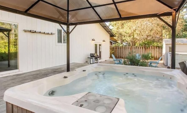 A hot tub under a covered patio with a wooden deck, a barbecue grill, and a wooden fence in the background.