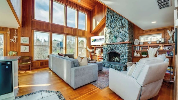 A spacious living room with a stone fireplace, wooden paneling, and large windows.
