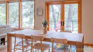 Ein Essbereich mit Holztisch und Stühlen, großen Fenstern mit Blick auf Bäume und einem kleinen Tisch mit Blumen in einer Vase.