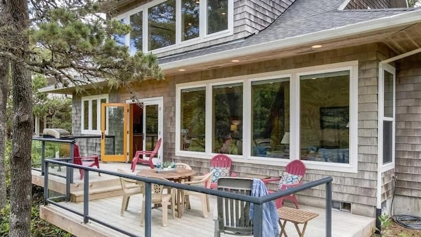Ein zweigeschossiges Haus mit einer Terrasse, großen Fenstern und Sitzgelegenheiten im Freien.
