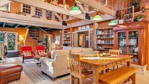 Amplia sala de estar con techo alto, vigas de madera, una mesa de comedor con fruta, un sofá y una estantería llena de libros y objetos decorativos.